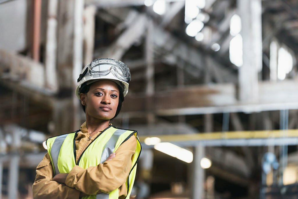 Portrait of a confident, young African American woman working in a manufacturing plant.  She is standing with arms folded, looking at the camera, in a storage facility or warehouse.  She is wearing a white hardhat and yellow safety vest.